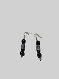 A pair of black beaded drop earrings with silver fishhook wires and white geometric carvings on the central beads, shown against a solid gray background.