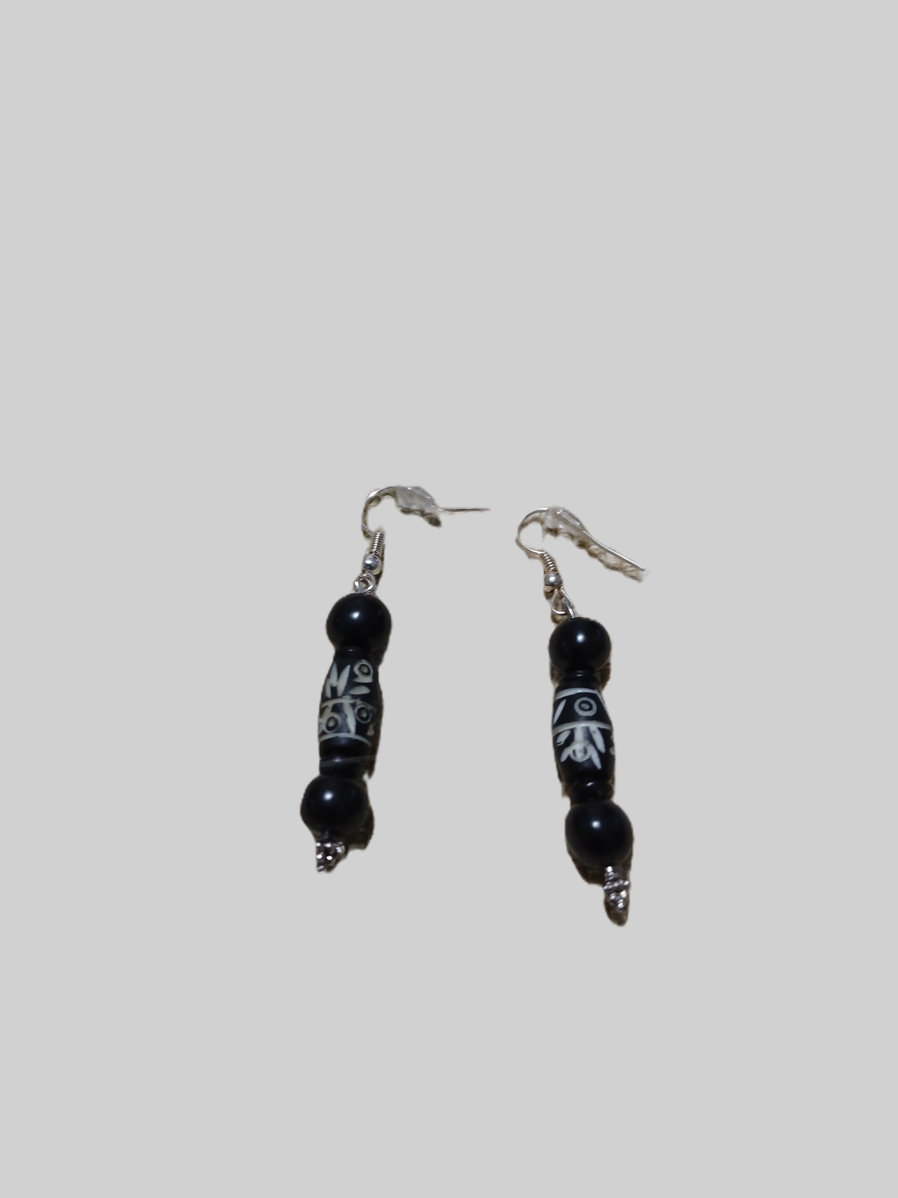 A pair of black beaded drop earrings with silver fishhook wires and white geometric carvings on the central beads, shown against a solid gray background.